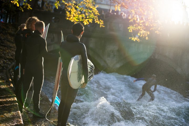 People wearing black wet suits and surfing on the Eisbachwelle. In the background is a bridge.