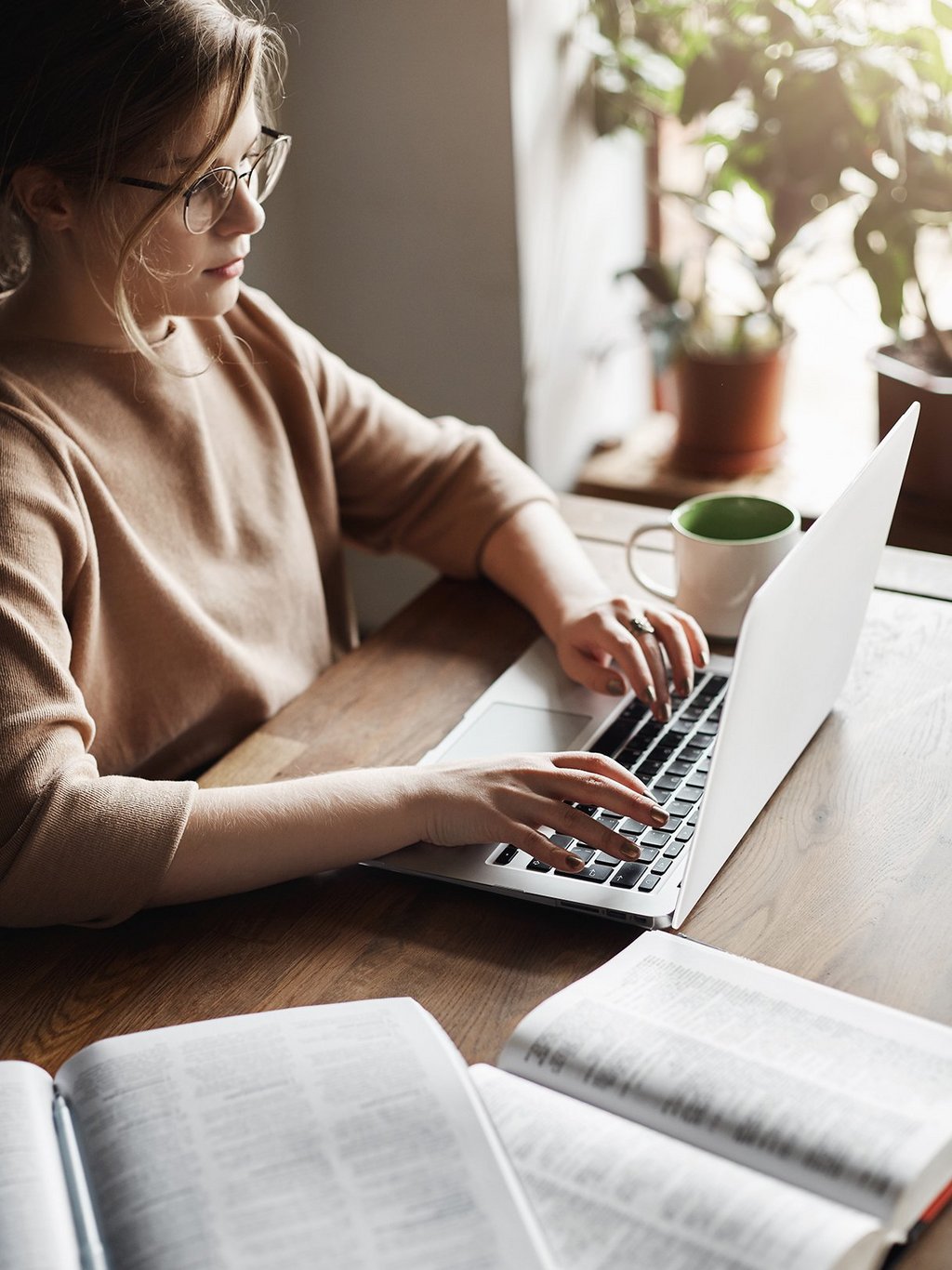 Vertical shot of determined and focused female student in glasses, sitting on wooden table, working with laptop and books, searching information 