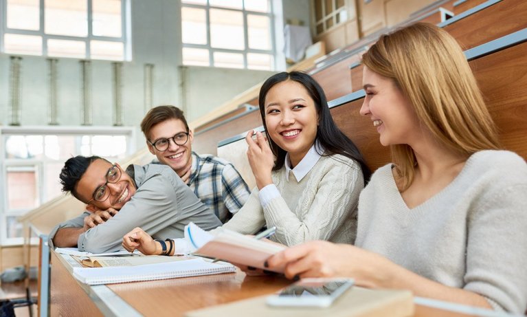 Multi-ethnic group of cheerful students sitting at desks in lecture hall and laughing, focus on two girls in front